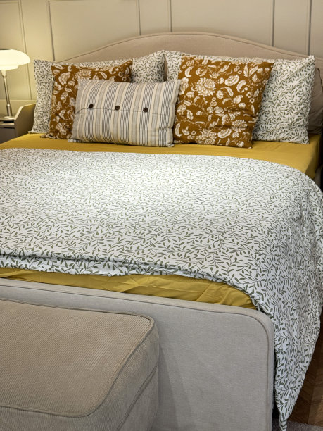 酒店房间双人床的特写图片，床头板上倚着一叠花图案的枕头，灰色和白色的折叠羽绒被，黄色的床单，床脚的卧室长凳，木质墙板，重点在前景图片下载