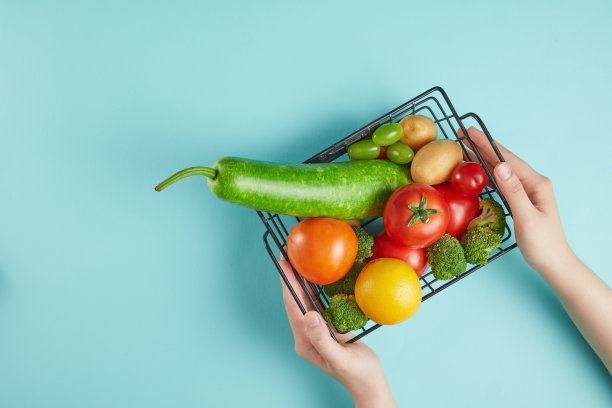 在蓝色背景的俯视图照片，美丽的手拿着一个黑色网格篮子的各种新鲜蔬菜放在桌子上。有机原产地产品广告图片，留白设计图片下载