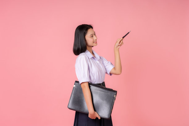 亚洲女高中生，短发，背着书包，穿着校服，用笔指着讲台。这张照片是在一个工作室用粉红色的背景拍摄的。图片下载