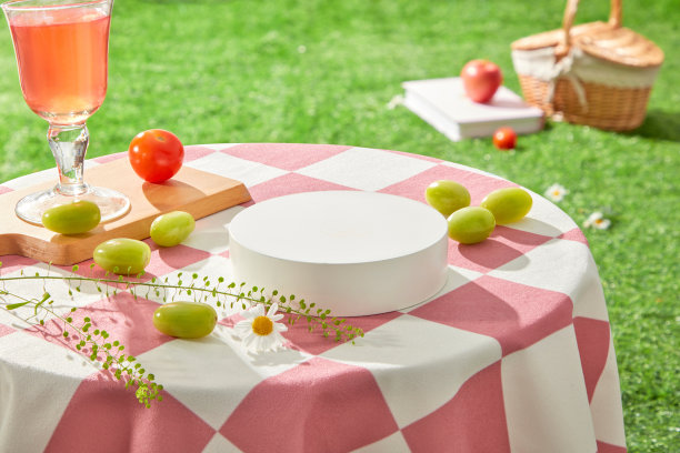 空白的讲台放在一张铺着粉红色格子桌布的桌子上，周围装饰着一些新鲜水果、一枝花和一杯红色液体鸡尾酒。高视角广告照片图片下载