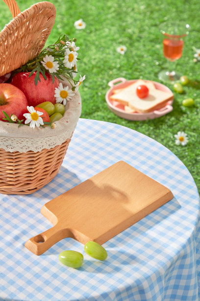 上面拍摄的户外野餐照片，一个空白的木托盘放在草地上的蓝色方格桌子上，旁边是一个装满水果的藤筐。为添加设计元素腾出空间图片下载