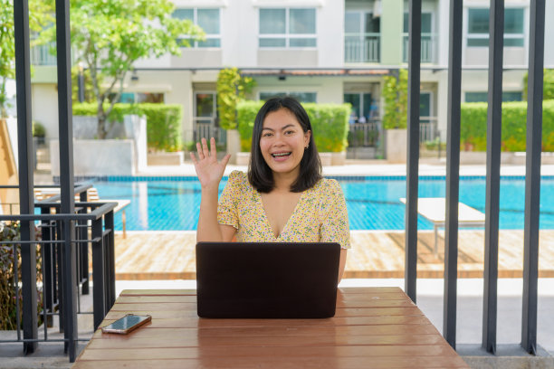 一名女子穿着黄色碎花连衣裙，在户外使用笔记本电脑，背景是游泳池图片下载