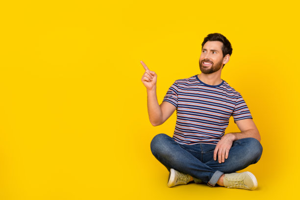 年轻人在休闲服装的全长照片坐在地板上点手指空的空间广告新的促销提供孤立的黄色背景图片下载