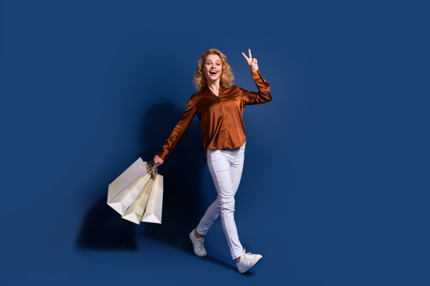 友好女孩的全尺寸照片，穿着丝绸衬衫牛仔裤，上面有v字符号，拿着购物袋，背景是深蓝色图片下载