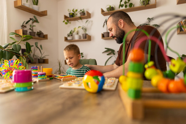 一位父亲和蹒跚学步的孩子在玩一场乱七八糟的字母游戏。深色头发和胡子的爸爸和浅色头发的男孩玩得很开心。他们坐在桌子旁，一边聊着幼儿园作业，一边玩得很开心。焊接时间。库存图片图片下载