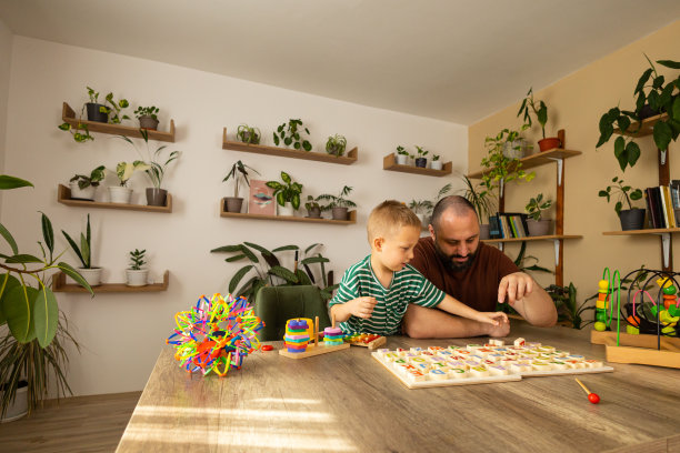 父亲和蹒跚学步的孩子正在玩一场乱七八糟的字母游戏。深色头发和胡子的爸爸和浅色头发的男孩玩得很开心。他们正坐在桌旁谈论幼儿园的作业。从远处拍摄。左拷贝空间。库存图片图片下载
