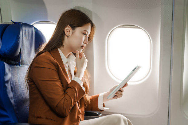 一位年轻的亚洲女商人穿着正装，坐在飞机座位上，拿着笔记本电脑和公文包，利用飞机上的Wi-Fi进行电话会议。图片下载