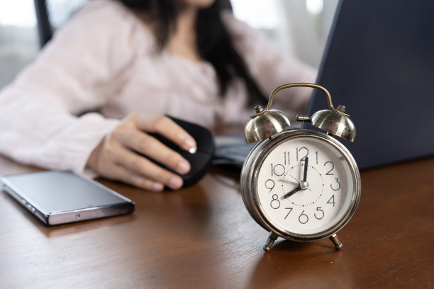 一位女士坐在办公桌前，手里拿着一台笔记本电脑和一部手机。桌子上放着一个大闹钟。图片下载