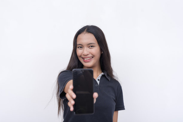 一位年轻的亚洲女性正在展示她手机上的一个应用程序。身穿深蓝色polo衫、卡其裤和白色运动鞋。手机应用程序布局与空白屏幕。白色背景的全身照片。图片下载