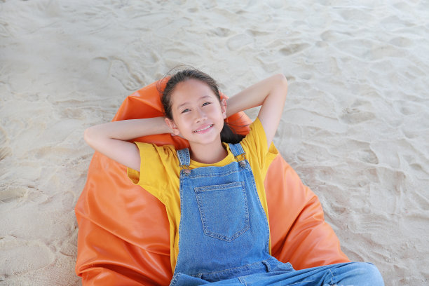 照片上的亚洲女孩微笑着穿着工装裤，看着镜头，在橙色的沙发床上放松，在沙滩上度假。图片下载
