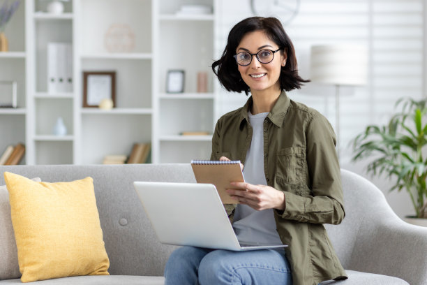 照片中，一个微笑的年轻女子戴着眼镜，穿着便服，坐在家里的沙发上，对着笔记本电脑，在笔记本上做着笔记，看着镜头图片下载