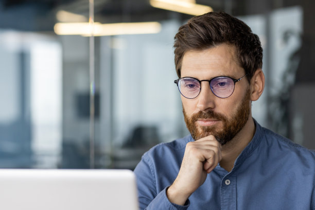 特写照片:一个戴着眼镜的深思熟虑的年轻人看着笔记本电脑屏幕，一只手捂着胡子，认真地思考着解决方案图片下载