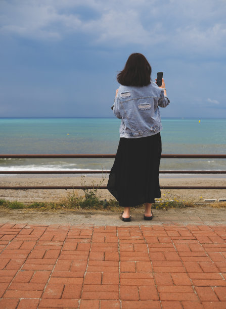 身穿黑色连衣裙的女子站在堤岸沙滩上，以海为背景拍摄照片图片下载