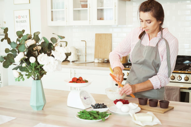 一个年轻的女人在厨房烤蛋糕，她穿着围裙站在柜台前，用搅拌器搅拌图片下载