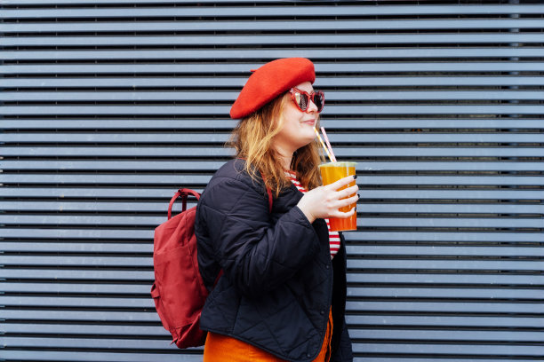 戴着心形太阳镜，穿着鲜艳衣服的大码女人微笑着，在条纹城墙旁喝着糖味的木薯奶茶。快乐的时尚时髦超重女孩。街头时尚。图片下载