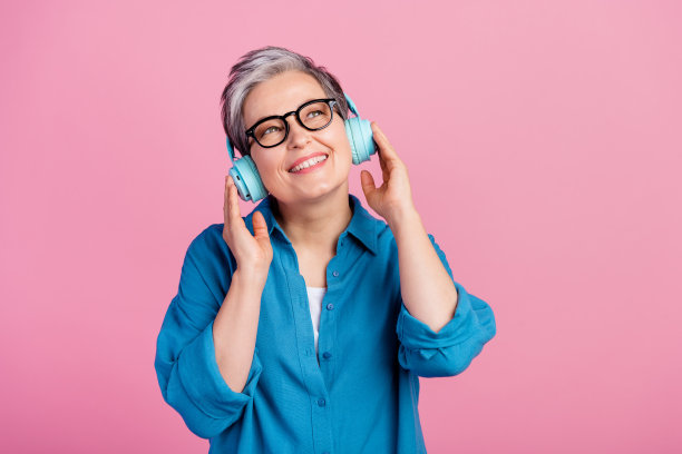 照片中有魅力的老年妇女听着音乐，戴着耳机，看起来空空荡荡，穿着时髦的蓝色衣服，孤立在粉红色的背景上图片下载