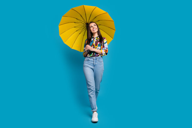 华丽的女人穿着印花衬衫牛仔裤的全长照片，拿着伞看销售空白空间孤立的蓝色背景图片下载