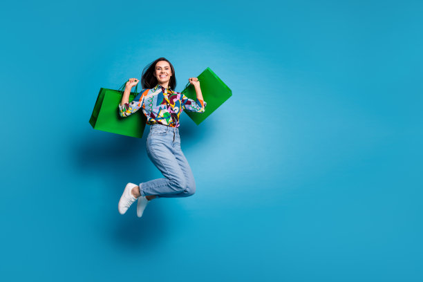 全长照片正面华丽可爱的女人穿印花衬衫牛仔裤抱着购物袋跳跃孤立的蓝色背景图片下载