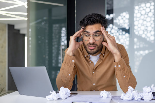 震惊和沮丧的年轻印度男子坐在办公桌前，带着笔记本电脑，皱巴巴地扔掉了纸，疲惫和沮丧地抱着头寻找新的想法图片下载