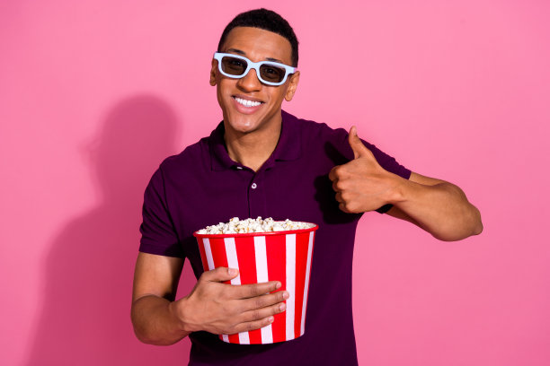 照片肖像漂亮的年轻男性3d眼镜拇指爆米花看电影穿着时尚的紫色服装孤立在粉红色的背景图片下载