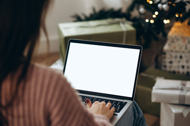 女人在笔记本电脑上工作，屏幕空白，有复制空间的模型。为了庆祝圣诞节，家里装饰了圣诞树和花环灯。图片下载