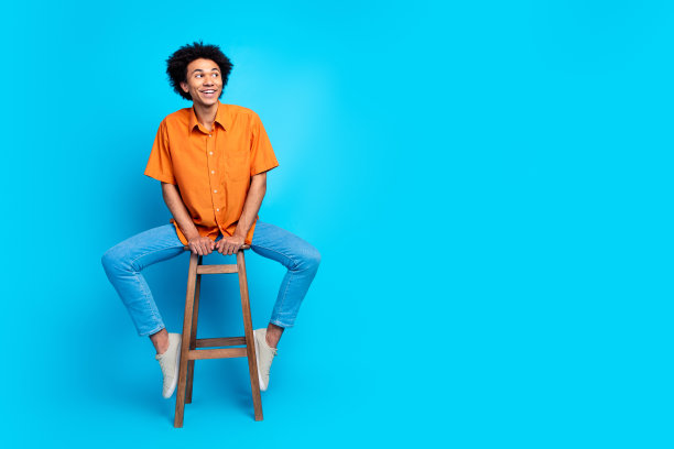 时髦酷的家伙穿着橙色衬衫坐在凳子上看空空间孤立的蓝色背景的全长照片图片下载