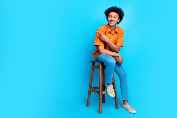 开朗积极的家伙穿着橙色衬衫的全长照片，坐在凳子上，显示空旷的空间孤立的蓝色背景图片下载