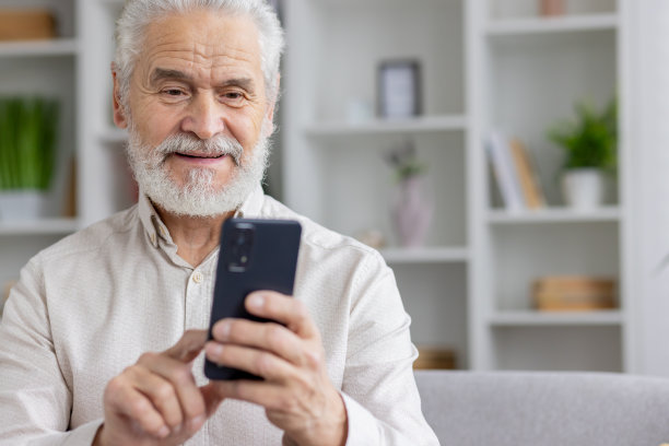 一个穿着白衬衫、头发花白、面带微笑的退休老爷爷的特写照片，他在家里玩智能手机图片下载