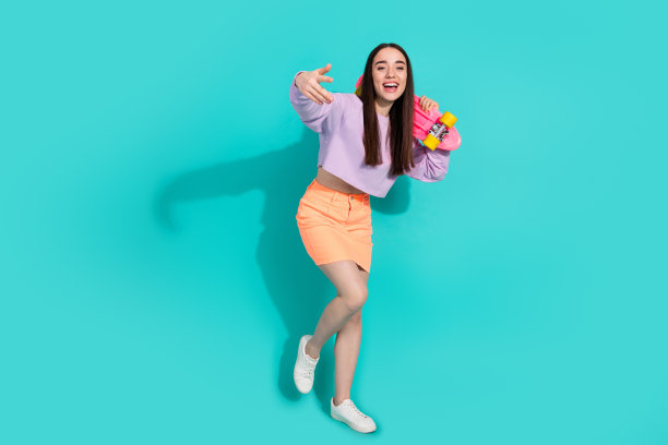 可爱的年轻女士手持滑板舞蹈的全身照片，穿着时尚的紫色服装，孤立于海蓝宝石色背景图片下载