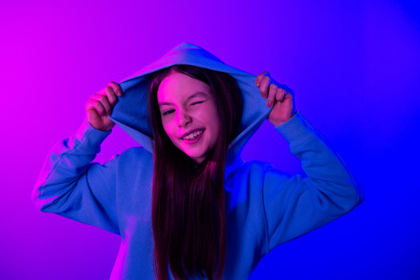 照片中开朗轻浮的小女士穿着运动衫手臂兜帽眨着眼睛空旷的空间孤立的霓虹紫色背景图片下载