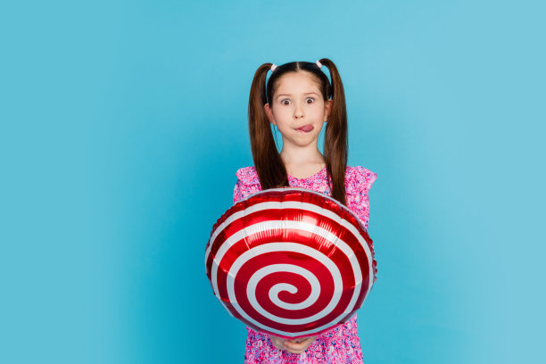 照片中开朗漂亮的小女士穿着粉红色的衣服舔着嘴唇，上升的棒棒糖气球空的空间孤立的蓝色背景图片下载