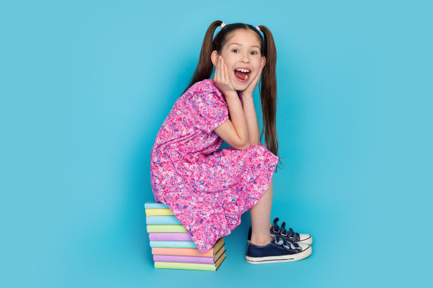 兴奋迷人的小女士的全身照片穿着粉红色的衣服坐在书堆空的空间孤立的蓝色背景图片下载