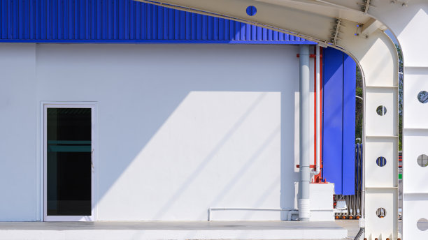 阳光和阴影投射在白色水泥和蓝色铝墙的工业办公楼上，曲线金属屋顶在停车场的前景上图片下载