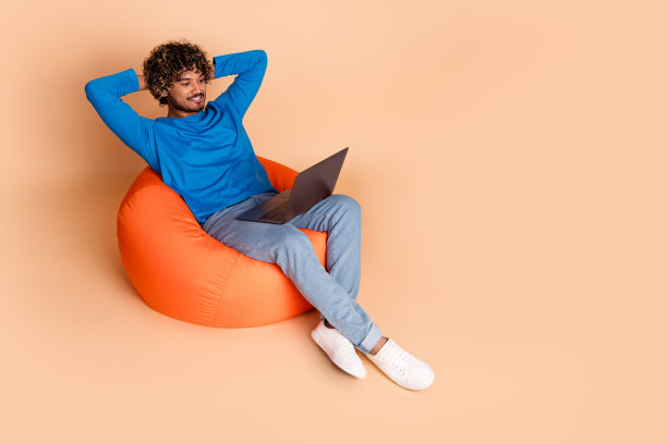 照片中开朗的好人穿着蓝色的衣服，坐着，抱着手臂在头后面，在米色的背景上空空荡荡的图片下载