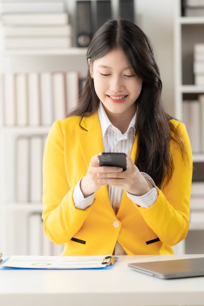 年轻的亚洲会计师在一个非正式的办公室里一边打电话，一边用笔记本电脑和移动应用程序工作。聊天，视频通话，业务联系和营销。在线的利润。垂直的图像。图片下载