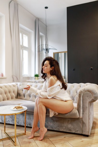 侧视图，漂亮的年轻女子闭着眼睛坐在灰色的沙发上，端着一杯茶靠近咖啡桌。穿着性感白衬衫的女士摆姿势图片下载