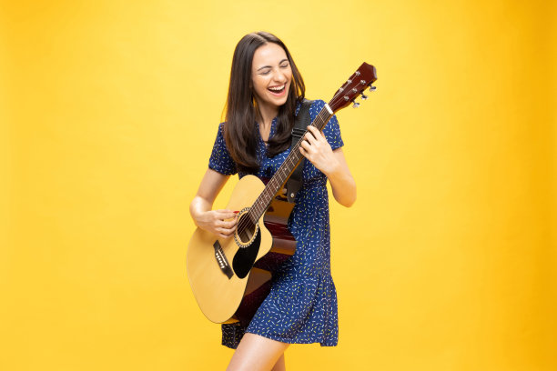 开朗可爱的女孩穿着时髦的衣服，拿着吉他乐器，在黄色背景照片上玩得很开心图片下载