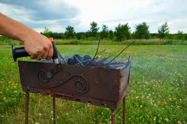 一名男子拿着带煤气罐的燃气灶，在烤架上点火的特写镜头。傍晚，大炮的火焰映在后院的绿草地上。图片下载