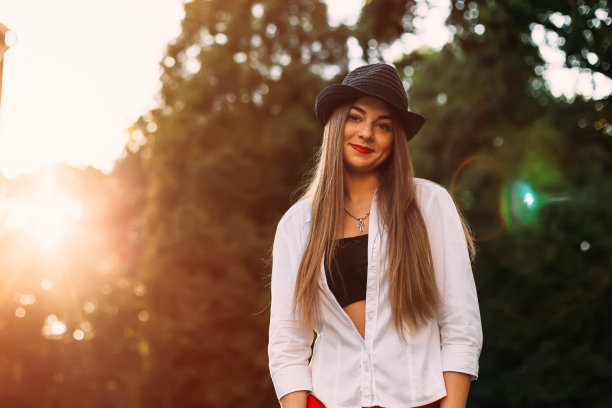 一个女孩化着漂亮的妆，穿着时髦的衣服，在绿树和阳光的背景下看着镜头。图片下载