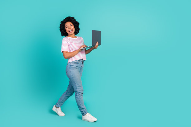 可爱的年轻女士拿着上网本走路的全身照片，穿着时尚的条纹衣服，孤立在海蓝宝石的背景上图片下载