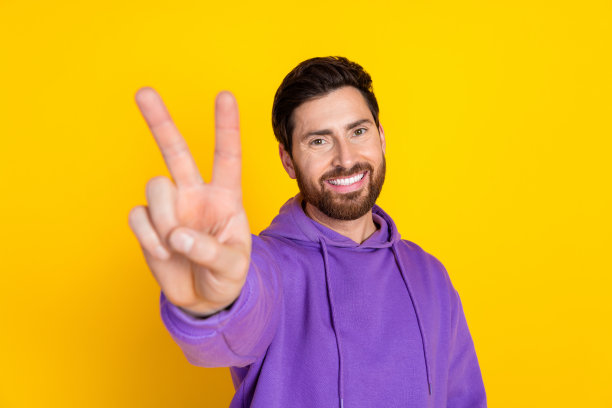 照片上，一个长着牙齿、满脸笑容的男人穿着紫色的卫衣，在黄色的背景上向你展示了v字符号图片下载