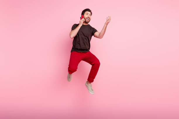 积极有趣的男人的全尺寸照片，穿着棕色t恤，在智能手机上说话，跳跃，在粉红色的背景上孤立的空间图片下载