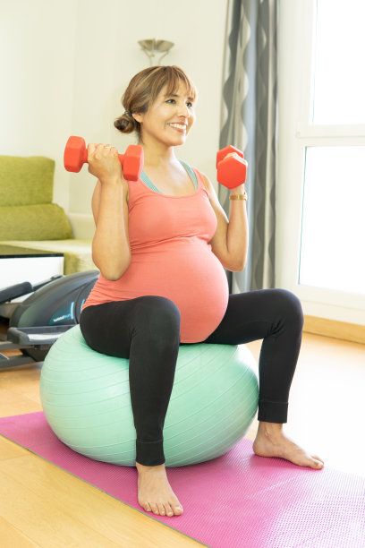 一位快乐微笑的孕妇坐在家庭健身房的瑜伽球上举着红色的杠铃。概念:产前锻炼，怀孕，婴儿保健。图片下载