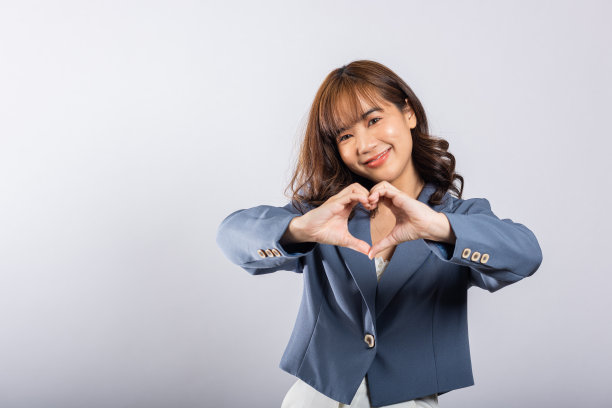 一位自信的商务女性穿着西装，微笑着在白色的背景上画出两只手的心形图案。一个美丽的年轻女性表达爱情的亚洲肖像图片下载