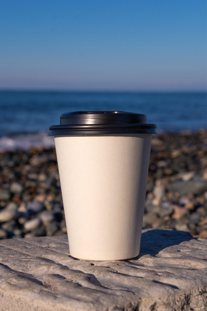 在阳光明媚的日子里，两个白色纸杯站在海边的一块石头上的特写，复制空间。外带咖啡图片下载