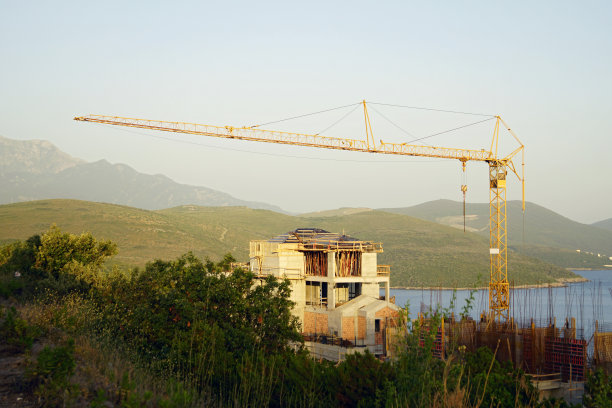 风景如画的建筑工地:未完工的混凝土房屋和高耸的塔式起重机映衬着群山和大海。图片下载