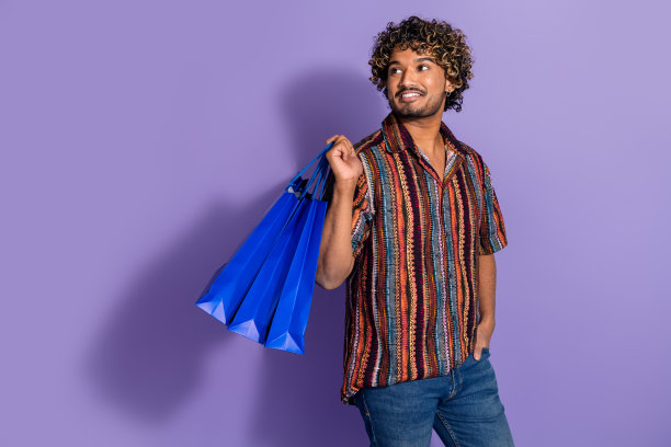照片上乐观的男人波浪的头发，穿着印花衬衫，看着销售空的空间，拿着购物袋孤立的紫色背景图片下载