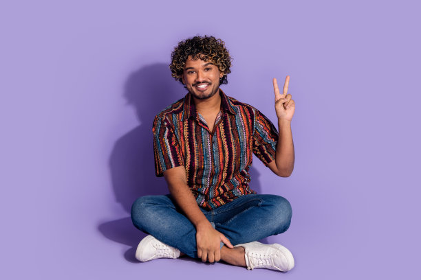 全尺寸照片的好心情的男人穿着印花衬衫牛仔裤裤子坐在显示v符号孤立的紫色背景图片下载