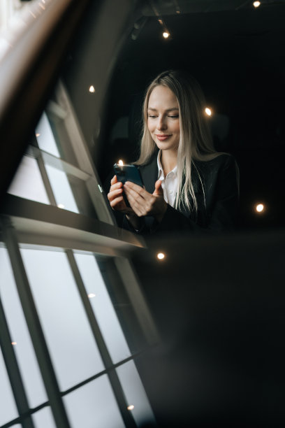 透过挡风玻璃的垂直照片，看到坐在汽车副驾驶座位上用智能手机阅读好消息的金发女郎。漂亮的年轻女子在浏览手机图片下载