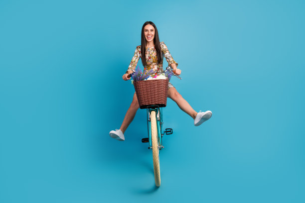 漂亮的年轻女孩骑自行车的全尺寸照片好玩的蔓延腿穿时髦的花朵印花服装孤立的蓝色背景图片下载